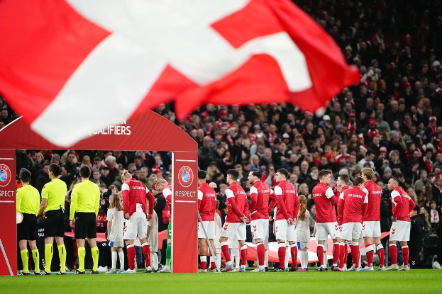 Gruppe von Menschen auf einem Fußballfeld mit einer roten und weißen Flagge im Vordergrund, einem Bogen mit der Aufschrift "Bayern Munich vs Bayern Munich Betting Tips & Preview" im Hintergrund und einer großen Stadionmenge.