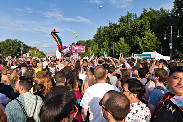 Eine große Menschenmenge steht vor einem Gebäude, einige halten Fahnen und Banner, mit einem Zelt, Laternenmasten, Bäumen und einem Luftballon im Hintergrund, versammelt zum Feiern der LGBT-Gemeinde.