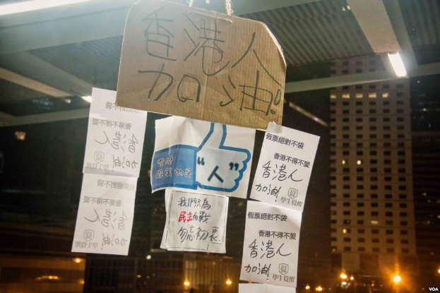 Eine Gruppe von Menschen vor einem Gebäude bei Nacht, mit beleuchteten Protestschildern an der Decke.