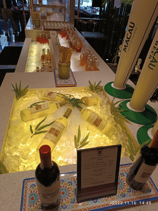 Ein Tisch mit Weinflaschen, Eiswürfeln, Pflanzen, Brettern und anderen Gegenständen beim Macau-Bierfestival, mit Menschen, Tischen, Stühlen und Glastüren im Hintergrund.