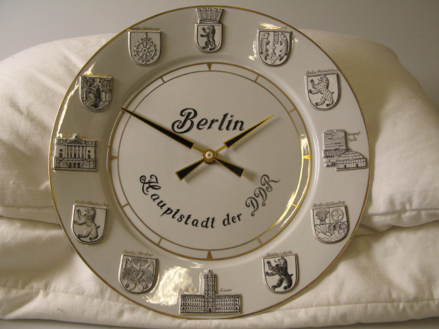 Eine Uhr mit dem Wort 'Berlin' darauf auf einem weißen Kissen gegen eine Wand gestellt.