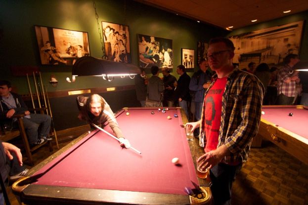 In einem Club spielen zwei Personen Snooker an einem von zwei Tischen, während mehrere andere im Hintergrund stehen und sich unterhalten, vor einer cremefarbenen Wand mit einigen Fotorahmen.