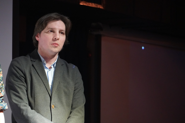 Zwei Personen nebeneinander auf einer Bühne, der Mann im Anzug und die Frau neben ihm, beide mit ernsten Gesichtern.