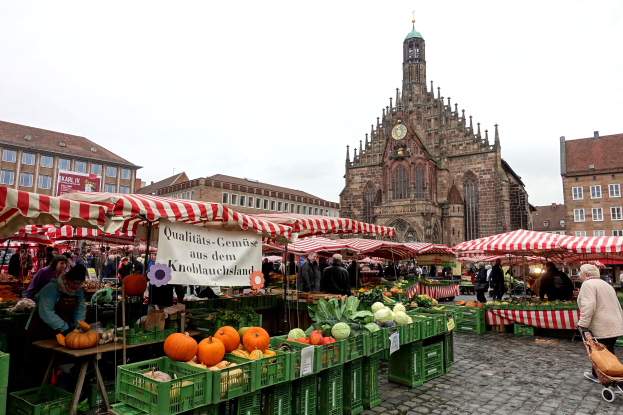 Ein belebter Markt in Nürnberg, Deutschland, mit verschiedenen Früchten und Gemüsen auf dem Stand, Menschen, die herumlaufen, Zelte, Gebäude mit Fenstern und ein Uhrenturm im Hintergrund unter einem sichtbaren Himmel.