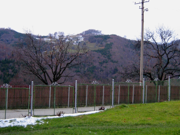 Eine grüne Landschaft mit einem Zaun, einem Pfahl und Bäumen, mit einigen Häusern im Hintergrund auf einem Hügel.