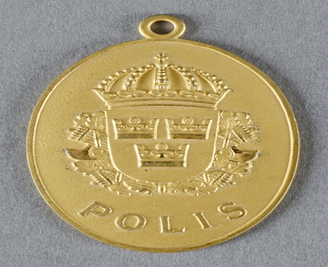 Goldene Medaille mit einer Krone, die wie ein Polizeibadge aussieht, in der Mitte des Bildes mit eingravierter Schrift.
