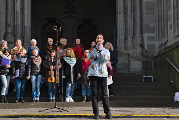 Eine Gruppe von Menschen, einige mit Musikinstrumenten, steht vor einem Gebäude mit Treppen und Geländern, während eine Frau in der Mitte in ein Mikrofon singt, umgeben von Mikrofonständern.