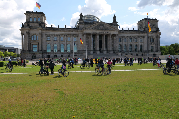 Eine Gruppe von Menschen, die Fahrräder vor dem Reichstaggebäude in Berlin fahren, das mit Fenstern, Säulen und Fahnen mit Stangen geschmückt ist, umgeben von Bäumen und Pflanzen, mit einem bewölkten Himmel und grasbewachsenem Boden.