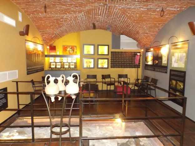 Ein Museumsausstellungsraum mit zahlreichen Tischen, Stühlen, Displaytafeln, gerahmten Gegenständen an den Wänden, Vasen, Beleuchtung und anderen Gegenständen.