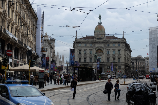 Eine belebte Stadtstraße mit einem geparkten Polizeiauto, Fußgängern mit Taschen, fahrenden Fahrzeugen, hohen Gebäuden, Bannern, Laternenmasten und Verkehrsampeln unter einem bewölkten Himmel.