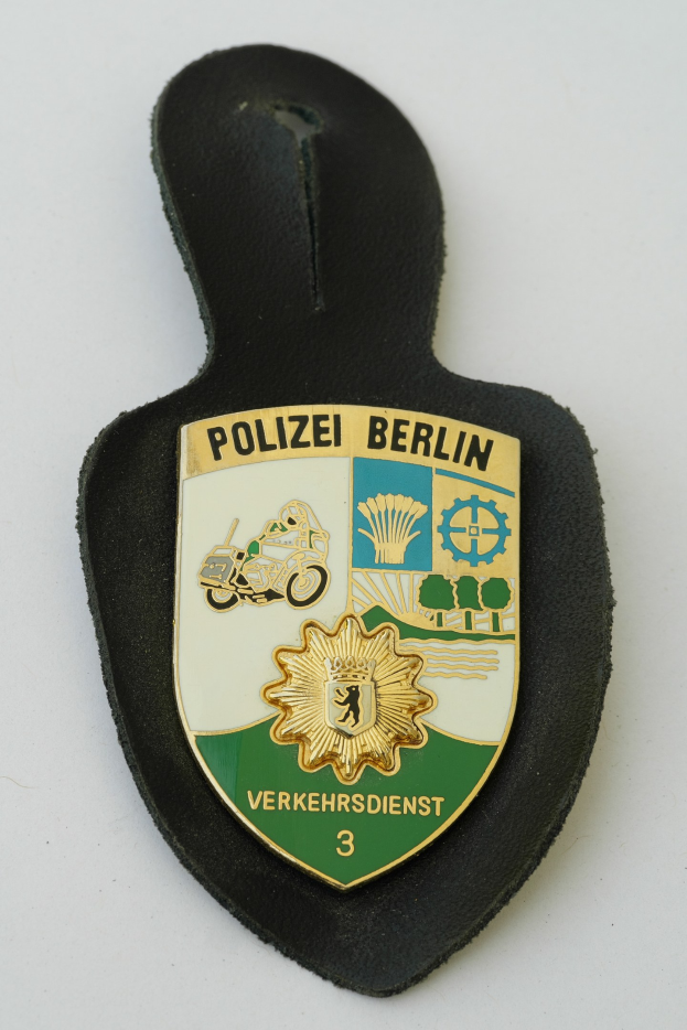 Ein schildförmiges Polizeibadgebnis mit "Polizei Berlin" in schwarzer Fettschrift auf einem blauen Hintergrund mit einem weißen Stern und Rahmen, das auf einer Oberfläche ruht.