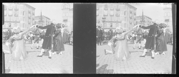 Ein Schwarz-Weiß-Foto einer Gruppe von Menschen, die auf der Straße tanzen, während eines Faschingsumzugs, mit Gebäuden, Straßenlaternen und Straßenlaternen im Hintergrund unter einem klaren Himmel.