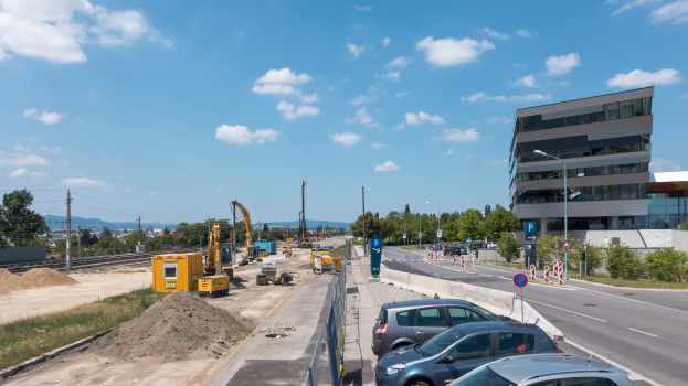 Eine Baustelle mit parkenden Autos, Gebäuden, Straßenlaternen, Straßenschildern, Absperrpoller, Bäumen, Pflanzen, Gras, Sand, Kränen, Bahnschienen, Strommasten mit Kabeln, Hügeln und einem bewölkten Himmel im Hintergrund.