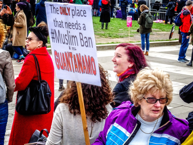 Eine Gruppe von Menschen steht auf Gras, einige halten Taschen, während eine Frau ein Schild hält, auf dem steht: "Der einzige Ort, an dem ein Muslimverbot benötigt wird, ist Guantanamo", vor einem Metallzaun, einem Schild, Pfählen, einem Gebäude mit Fenstern und Bäumen.