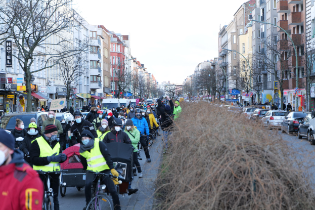 Eine große Gruppe von Menschen mit Masken und Sicherheitswesten auf Fahrrädern die eine von Bäumen gesäumte Straße entlangfahren, mit Gebäuden, Laternenmästen und Texttafeln, begleitet von Fahrzeugen und trockenem Gras unter einem klaren blauen Himmel.