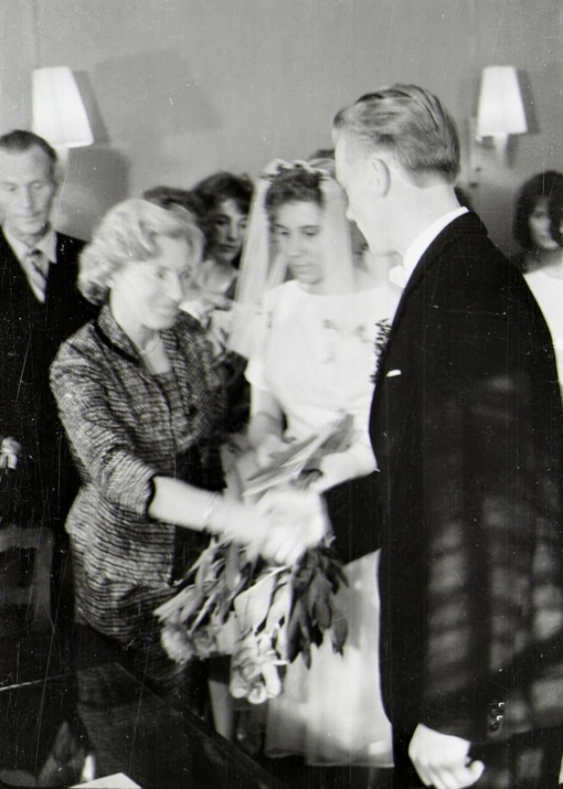 Ein Schwarz-Weiß-Foto eines Brautpaares bei der Trauung, das sich die Ringe ansteckt, wobei die Braut in einem weißen Kleid und Schleier mit einem Strauß Blumen in der Hand zu sehen ist und der Bräutigam in einem Anzug und Krawatte steht, mit Wandlampen im Hintergrund.