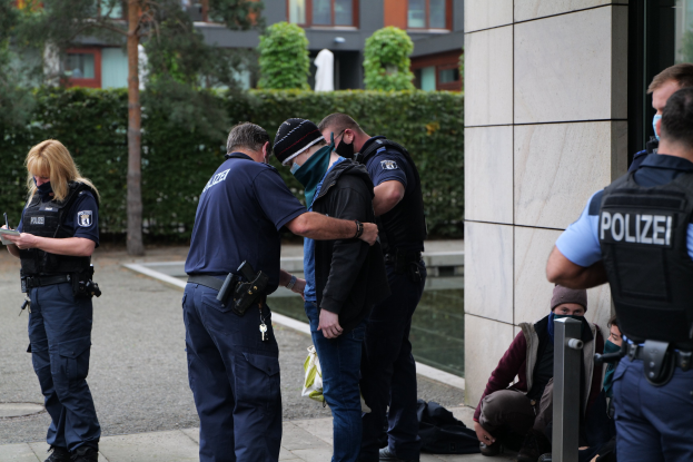 Eine Gruppe von Polizeibeamten vor einem Gebäude stehend, mit einer Frau, die ein Buch und einen Stift hält, und mehreren Menschen in der Nähe sitzend und stehend, vor dem Hintergrund von Pflanzen, Bäumen und Gebäuden.