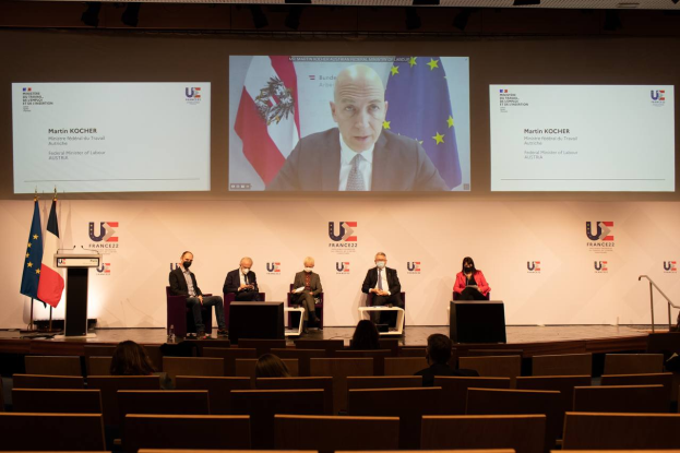 Eine Gruppe von Menschen sitzt auf einer Bühne in einem halben Kreis um ein zentrales Podium mit Mikrofon, flankiert von Flaggen, mit einem Bildschirm im Hintergrund, der Text und Bilder anzeigt, während einer Pressekonferenz der Europäischen Union.