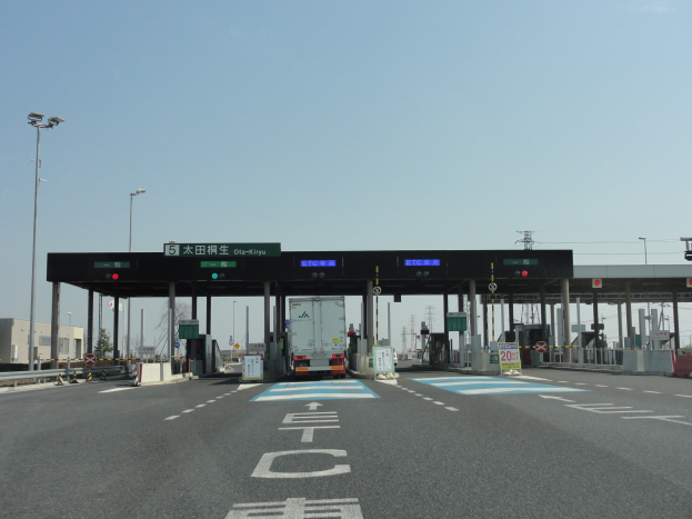 Eine Mautstation mit einem davor geparkten Lkw, Straßenrandmarkierungen in Weiß und Blau, Laternenmasten, Schilder, Gebäude, Bäume und einen klaren blauen Himmel.