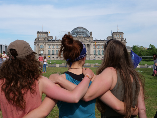 Drei Frauen in bunten Kleidern stehen vor dem Reichstag in Berlin, mit einer Menge, Fahnen und Bäumen im Hintergrund.