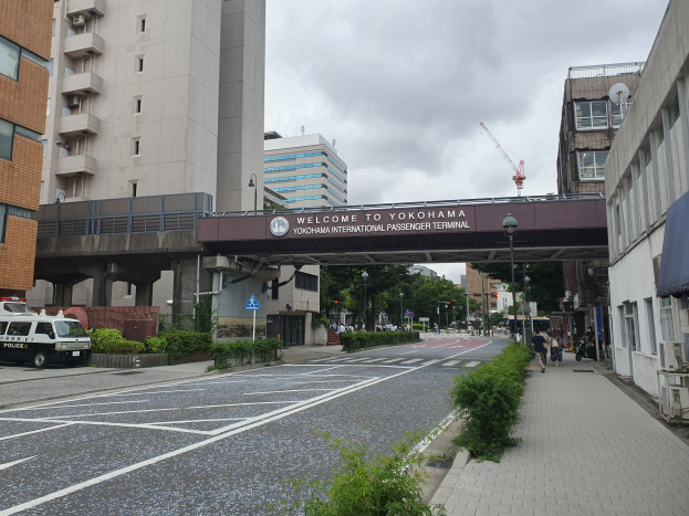 Eine Stadtstraßenszene mit einem geparkten Polizeiwagen, Gebäuden, Straßenlaternen, Straßenschildern, Kraftfahrzeugen, Fußgängern, Bäumen und einem bewölkten Himmel am Yokohama International Passenger Terminal.