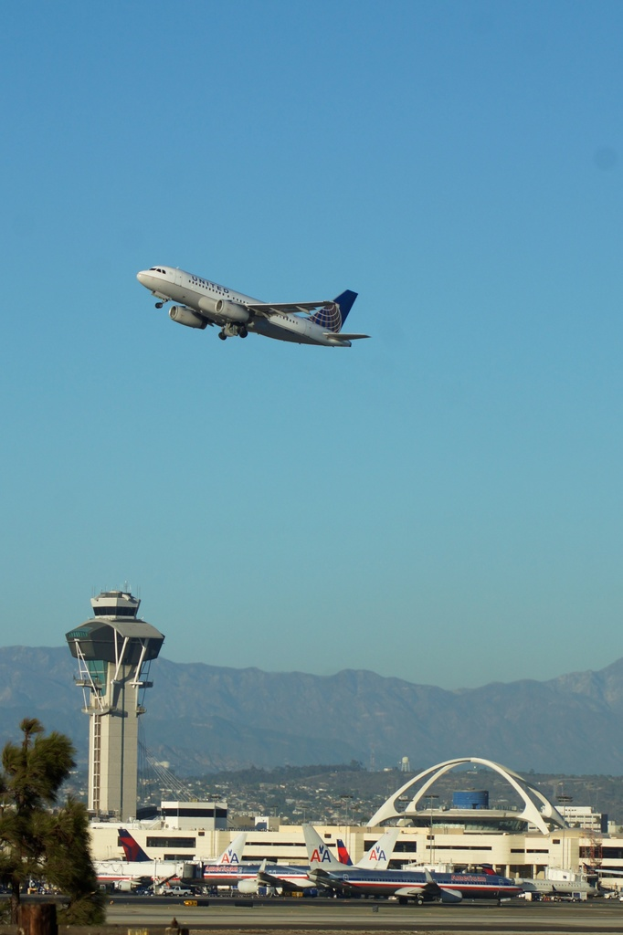 Ein Flughafenblick mit einem weißen Kontrollturm im Vordergrund, parkenden Flugzeugen am Boden, entfernten Gebäuden und einem Flugzeug am Himmel.