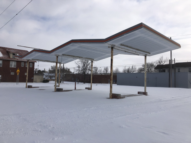 Eine Tankstelle steht in der Mitte einer verschneiten Straße, umgeben von Gebäuden, Bäumen, Polen, Drähten und Schildern, unter einem bewölkten Himmel.