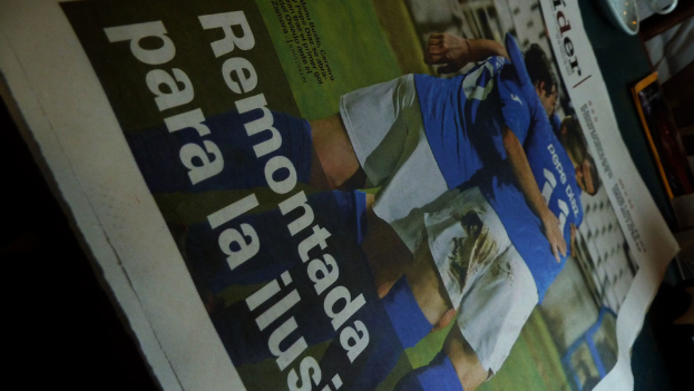 Eine Zeitung auf einem Tisch zeigt drei Männer in blauen Hemden, möglicherweise beim Fußballspielen, mit Text am unteren Rand des Bildes.