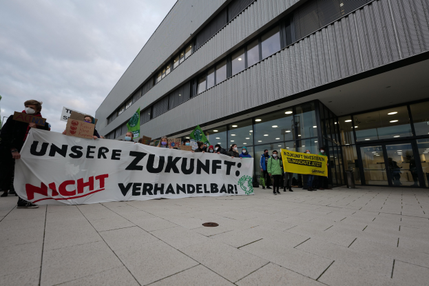 Gruppe von Menschen, die eine Banner mit der Aufschrift "Unsere Zukunft Verhandelbar" vor einem Glasfront-Gebäude bei Nacht halten, mit einem bewölkten Himmel im Hintergrund.