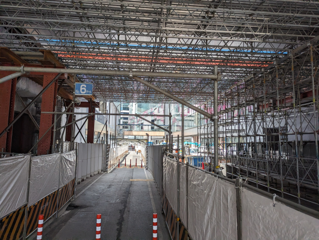 Baustelle mit umfangreicher Gerüstkonstruktion, einem Zaun, einem Schild, Metallrahmen, benachbarten Gebäuden und einem bewölkten Himmel.