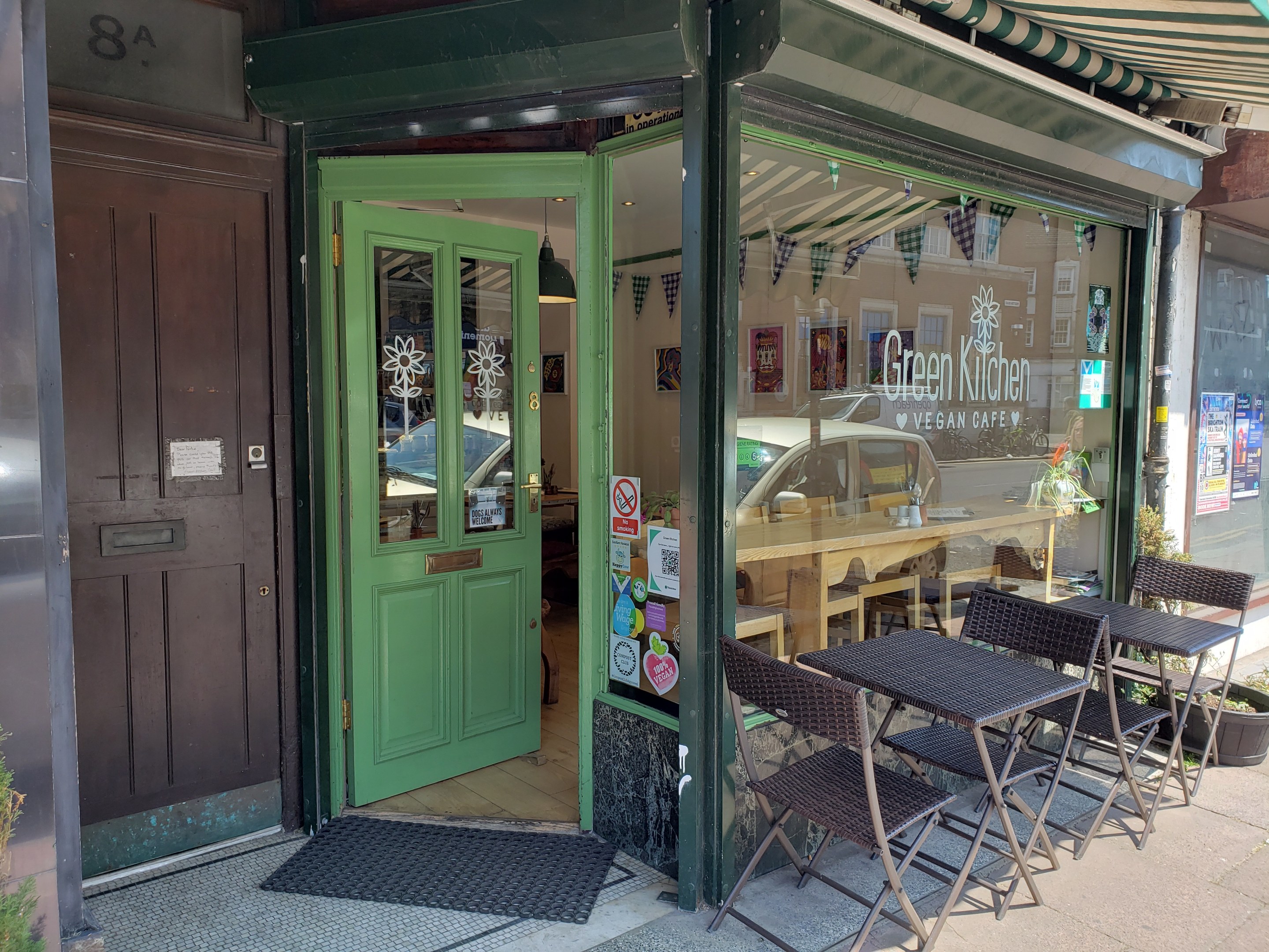 Außensitzbereich des Green Kitchen Vegan Cafe mit Tischen, Stühlen, Pflanzen, einer Matte und einer Glastür mit Postern, der in einen Innenbereich mit Tischen, Stühlen und Deckenleuchten führt.