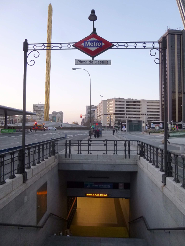 Eine belebte Stadtkulisse mit Geb├Ąuden, Fahrzeugen, Passanten, einem U-Bahn-Eingang und einem Schild ├╝ber ihm.