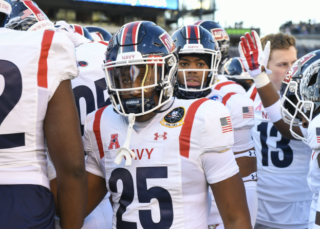 Eine Gruppe von Männern in Football-Uniformen steht auf einem Feld mit Helmen und Handschuhen, mit einer unscharfen Menge, einem Scoreboard mit der Aufschrift "Auburn vs Virginia Tech - 11/28/15 College Football Pick, Odds, and Prediction" und Himmel im Hintergrund.