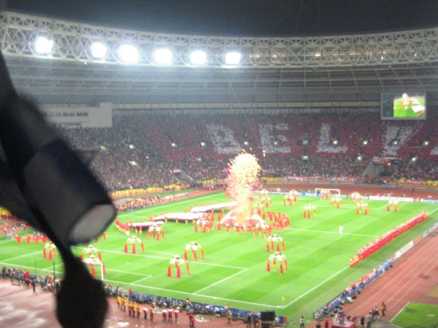Ein Stadion mit Flutlicht, Eisengittern, Zuschauern, stehenden Personen auf dem Boden, Werbetafeln, Anzeigetafeln, einer Überwachungskamera, Fahnen und Deko.