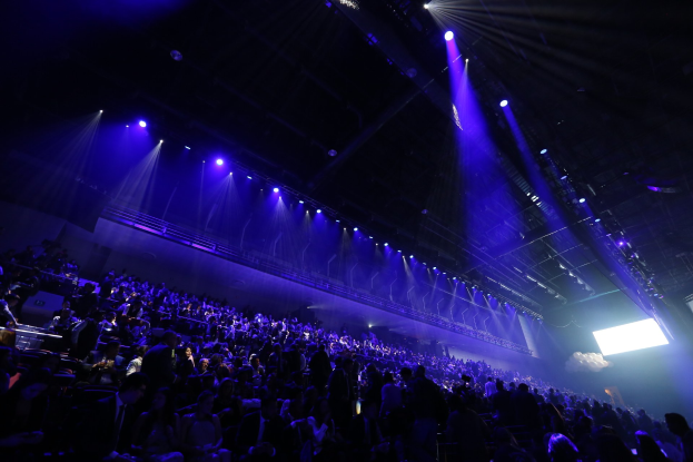 Große Menschenmenge vor einer Bühne mit Deckenbeleuchtung, rechte Seite zeigt einen Bildschirm in einer Konzerthalle.