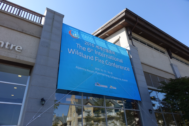 Ein modernes Gebäude mit Glaswänden und Lichtern, das ein Banner mit Text trägt, während der 6. Internationalen Wildland-Feuer-Konferenz in Pyongyang, Nordkorea, mit einem Baum auf der rechten Seite und dem Himmel oben sichtbar