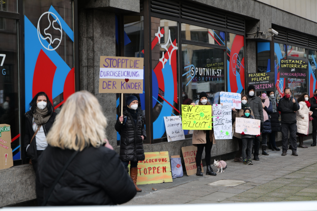 Eine Gruppe maskierter Personen mit Schildern vor einem Glasgebäude protestierend, mit Kameras, Texttafeln und einem Hund sichtbar.