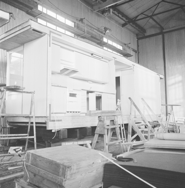 Ein Schwarz-Weiß-Foto eines im Bau befindlichen Gebäudes mit Leitern, Holzboxen und anderen Gegenständen, die auf dem Boden verstreut sind, und einer Wand mit Fenstern und Lichtern im Hintergrund.