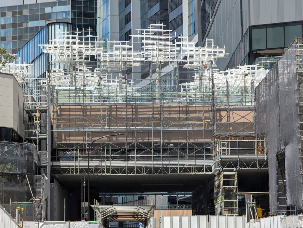 Großes Gebäude mit Gerüst, umgeben von Bäumen links und einer Straße unten.