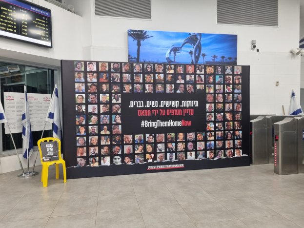 Eine große Wand mit mehreren Fotos, ein gelber Stuhl, israelische Regierungsflaggen auf Stangen, eine Tafel mit der Aufschrift "Bring Them Home Now", ein montierter Bildschirm, Deckenleuchten und zusätzliche Gegenstände.