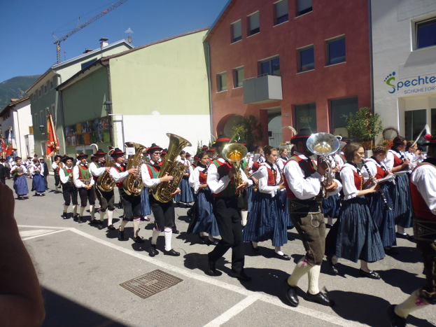 Eine Gruppe von Menschen in traditioneller bayrischer Tracht, die auf einer Straße mit Gebäuden, einigen Fahnen haltend, spielen und ein Hügel und ein klarer blauer Himmel im Hintergrund zu sehen sind.