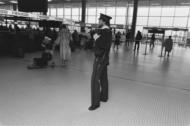 Ein Polizist in Uniform steht in einem Flughafen-Terminal und hält eine Waffe in der Hand, umgeben von Menschen und Gepäckwagen, mit Pfosten, Texttafeln, Deckenlampen und Glaswänden im Hintergrund.