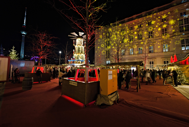 Ein belebter Weihnachtsmarkt in Berlin, Deutschland mit Menschen um dekorierte Stände, Lichter, Bäume, Gebäude, Laternenmasten und einen Turm unter einem dunklen Himmel.