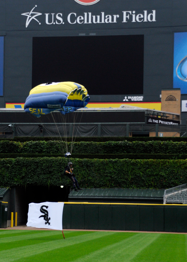 Eine Person gleitet mit einem Gleitschirm über ein Baseballfeld, umgeben von grünem Gras, Pflanzen und einem Banner mit Text, mit Tafeln mit Text und Logos und einem Gebäude im Hintergrund.