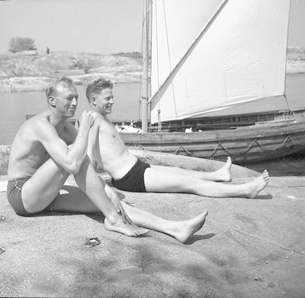 Zwei Männer in Badesachen sitzen am Strand neben einem Segelboot, mit blauem Wasser, grünen Bäumen und einem hellen Himmel im Hintergrund.