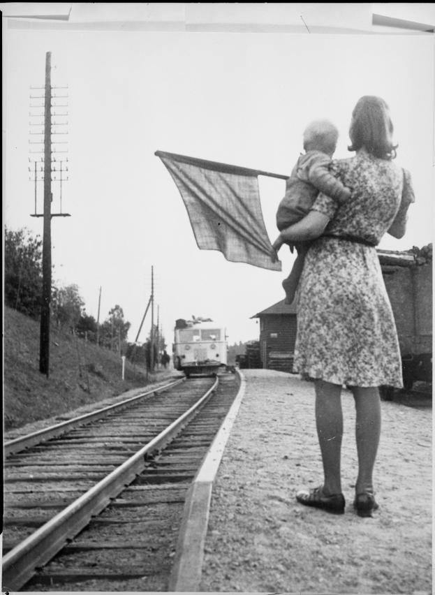 Eine Frau in einem Kleid und Schuhen steht auf den Bahnschienen und hält ein Baby, während ein Zug im Hintergrund näherkommt; Pfähle und Bäume sind links zu sehen und der Himmel ist oben zu erkennen.