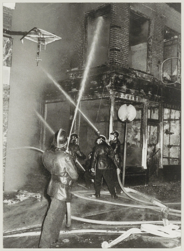 Eine Gruppe von Feuerwehrmännern in Helmen, die Schläuche halten, steht vor einem brennenden Gebäude, in Schwarz-Weiß abgebildet.