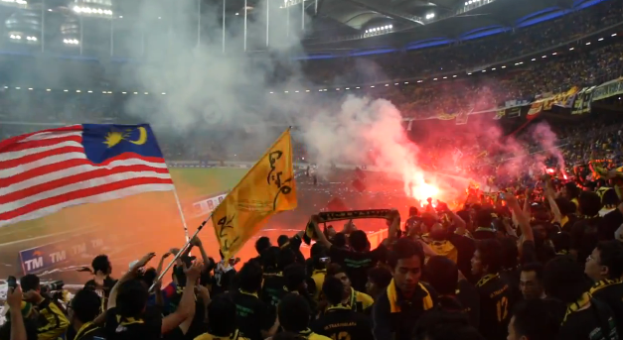Eine gro├če Menge Menschen in einem Stadion mit Bengalos, Fahnen und einer Kamera, mit Rauch und Schildern mit der Aufschrift "Malaysia vs Malaysia Live Streaming" im Hintergrund.