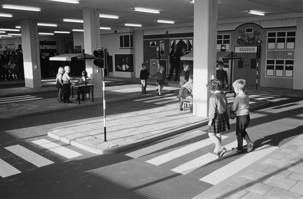 Schwarz-weißes Foto von Kindern, die eine Straße an einer Ampel überqueren, mit Fußgängern in der Nähe, einer Verkehrsampel im Vordergrund und Säulen mit Deckenleuchten im Hintergrund.