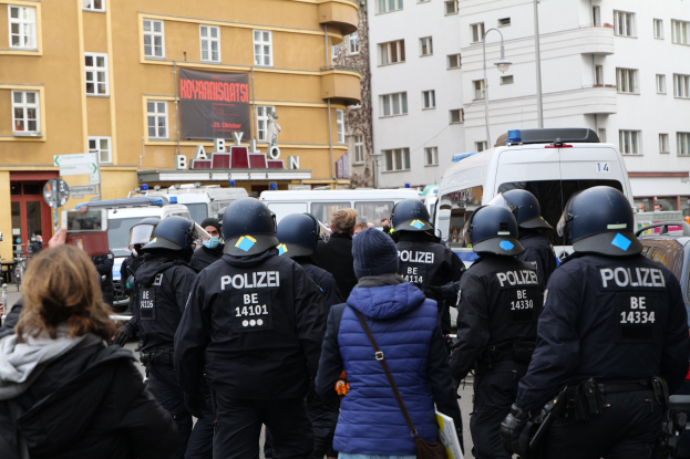 Polizeibeamte in Uniform vor einer Menge von Demonstranten, einige mit Helmen und Jacken, mit Fahrzeugen, Gebäuden, Laternen und einem Banner im Hintergrund während einer Demonstration in Berlin, Deutschland.